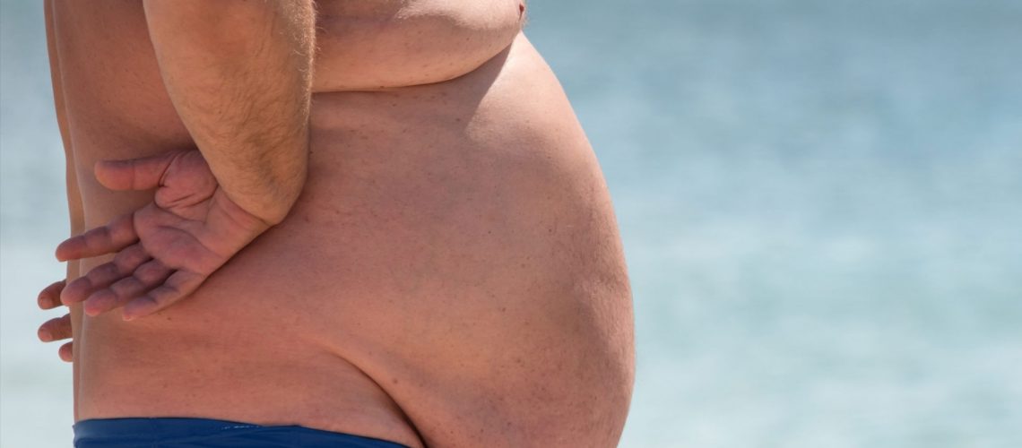 obese man on beach in small bathing suit with giant belly hanging in front